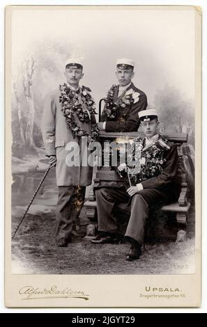 Ateljé portrait - three men dressed in student caps, with cane and garnished with flowers. One sits on bench, the other two stands, Uppsala 1899. The picture taken in 1899 by Alfred Dahlgren in Ateljé on Dragarbrunnsgatan 48, Uppsala. The men in the picture can be identified in pictures included in a collection of photos donated by Kerstin Hallström, Uppsala, 1976. These pictures are linked to Father Harald Falk's student time (1895-1905) in Uppsala. 1899 - 1899historic event, name related to Objek: Falk, Harald Stock Photo