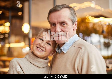 Smiling beautiful mature couple in beige sweaters standing in modern restaurant and looking at camera Stock Photo