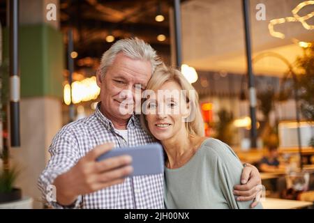 Handsome senior man with wrinkles embracing wife while posing with her for selfie on phone Stock Photo