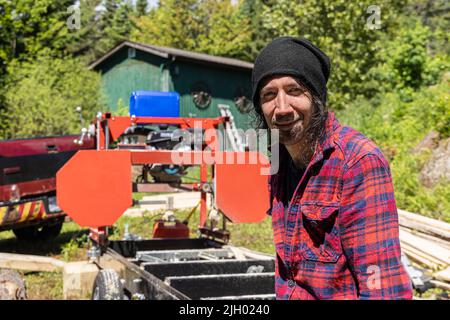 Close up portrait of a male lumberjack in mid thirties at work outdoors. Blurry machinery and cabin is seen in background with copy space to left. Stock Photo