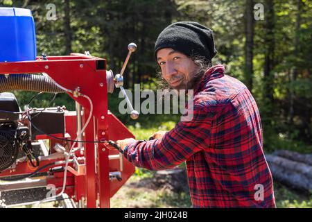 Portrait of a sawmill machine operative at work with black hair and stubble wearing a red checkered shirt, using a mobile saw trailer with copy space. Stock Photo