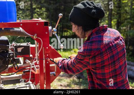Lumberjack is seen from behind wearing black hat and red checked shirt, operating an industrial saw in woodland with blurry trees in background. Stock Photo