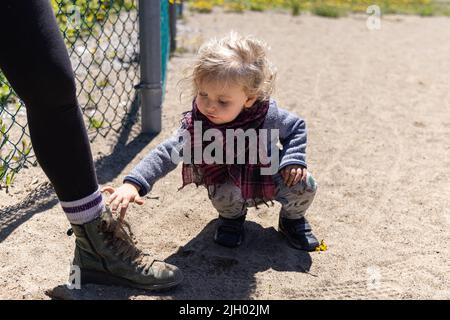 Close up shot of a crouching two year curious boy, wearing red scarf, throwing sand into parents laced up boot on a sandy playground. With copy space. Stock Photo