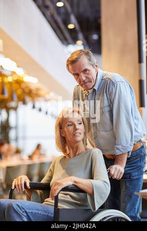 Portrait of content careful senior man in denim outfit pushing wheelchair with beautiful wife in cafe Stock Photo