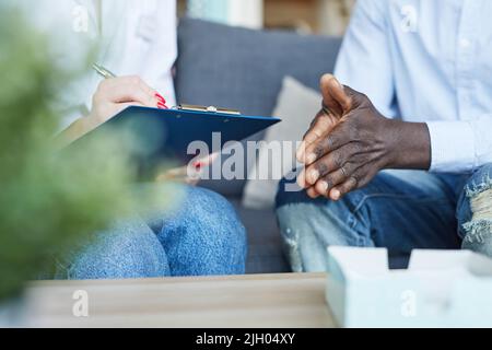 Close-up of unrecognizable psychologist sitting on sofa and making notes about patients problem while talking to black man at therapy session Stock Photo