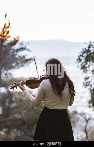 Brunette woman seen from back playing violin outside in the mountain. Vertical Stock Photo