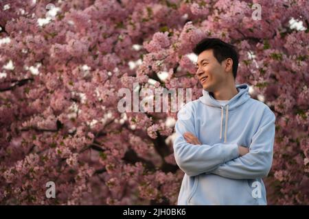 smiling Asian young man arm crossing, side face looking away. Blur pink sakura tree background Stock Photo