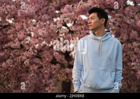cool handsome Asian young man hands in pockets, side face looking away. Blur pink sakura tree background Stock Photo