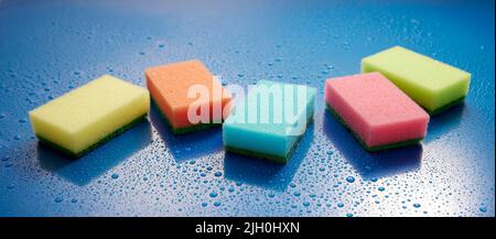 Collection of multicolored wash sponges placed on wet blue background covered with drops of water in light room during household routine Stock Photo