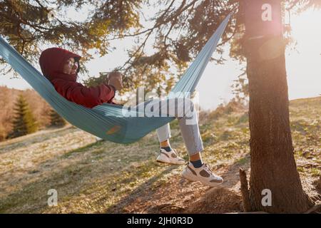 Woman in glasses and hood lying in hammock with thermos and enjoying wild nature in forest. Stock Photo