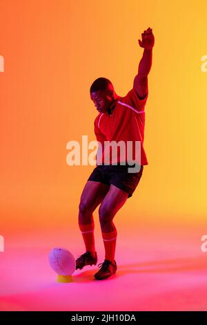 African american male rugby player kicking rugby ball over pink lighting Stock Photo