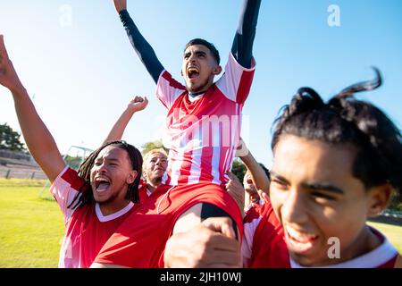 Multiracial male players carrying cheerful teammate on shoulders screaming while celebrating goal Stock Photo