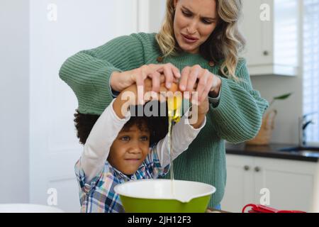 Multiracial mother making face while assisting son with afro hair in breaking egg in bowl in kitchen Stock Photo