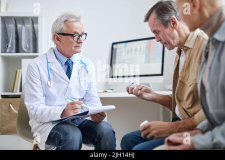 Serious senior male doctor in glasses making notes in clipboard while prescribing medication for mature patients Stock Photo