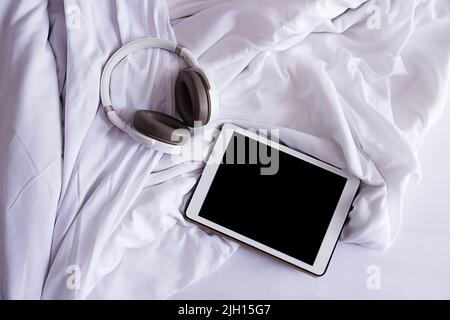 a tablet and headphones lie on a crumpled white bed, top view, close-up Stock Photo