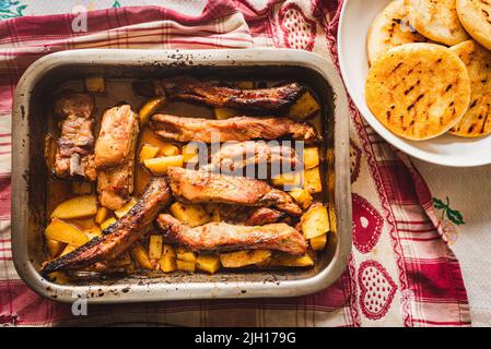 grilled pork ribs on table inside oven aluminum container with venezuelan arepas and potatoes, table cloth, and warm natural light Stock Photo