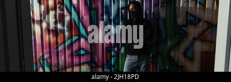 African american hooligan in hoodie and face mask standing near graffiti on wall, banner Stock Photo