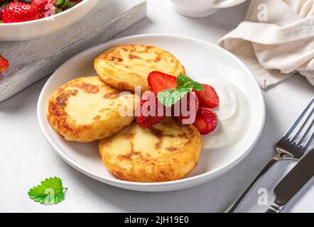 Cottage cheese pancakes with strawberries, mint and sour cream in a white plate on a gray background. Stock Photo