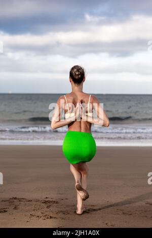 Anonymous female in skirt and bra clasping hands behind back while doing Garudasana pose on sandy beach on stormy day Stock Photo