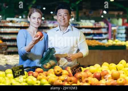 Portrait of young multi-ethnic family, Asian man and woman in supermarket shopping, smiling and looking at camera, couple chooses apple fruit Stock Photo
