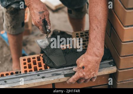 Worker or bricklayer works with trowel laying bricks. Builder makes brickwork on construction site, close up on hands. Stock Photo