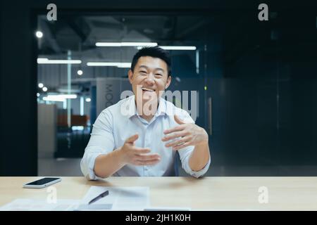 Online business training. Portrait of an active young Asian businessman. Conducts online business training on camera, sits at a table in a modern office, talks, explains, waves his hands, smiles Stock Photo