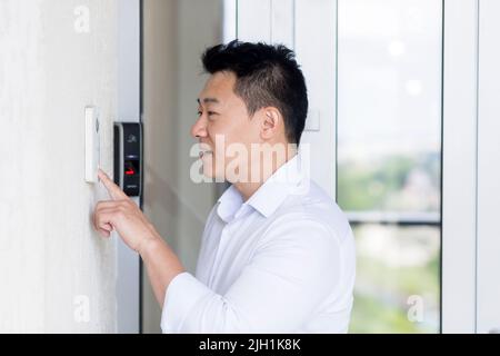 Close-up photo. Portrait of a young handsome Asian man, calling the intercom of the house, pressing the button, waiting for the door to open. Stock Photo