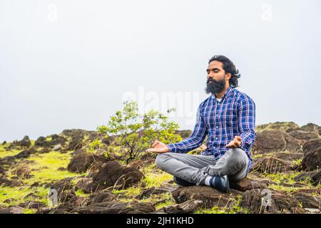 Young beard man with eyes closed doing meditation or yoga on top of hill - concept of self caring, health care and wellness Stock Photo