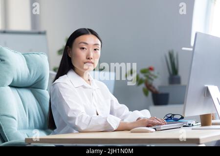 Close-up photo. Portrait of a young beautiful Asian woman secretary sitting at the desk at the computer in the office in business clothes, looking at the camera. Stock Photo