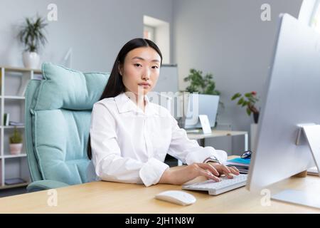 Close-up photo. Portrait of a young beautiful Asian woman secretary sitting at the desk at the computer in the office in business clothes, looking at the camera. Stock Photo