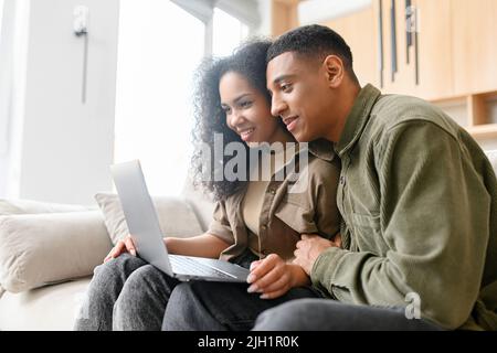 Young multiracial couple using laptop while sitting at the sofa at home. Spouses watching movie on laptop. Happy man and woman embracing while relaxing on couch Stock Photo