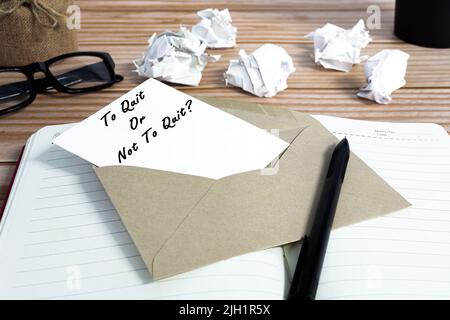 To quit or not to quit written on notepad with trash paper background on wooden desk. Decision making to quit or leave job. Employment and resignation Stock Photo