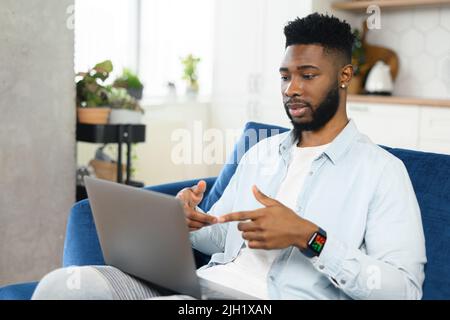 Serious concentrated male student involved virtual meeting, webinar. Black man making video call on the laptop, sitting on the sofa at home, talking and gesturing, explaining something Stock Photo