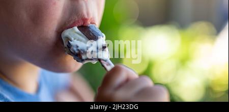 young boy eating an ice-cream bar with a chocolate coating outside on a hot day.  Stock Photo