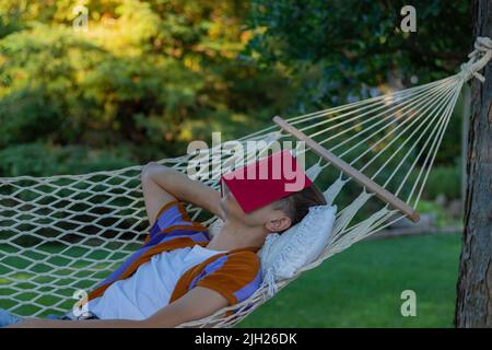 Young man resting and sleeping in a hammock in spring time with a red book covering his eyes and face in the garden Stock Photo