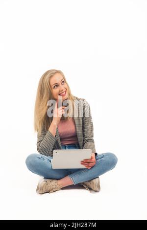 Gorgeous blond European young adult woman in jeans and checked blazer sitting cross-legged with her laptop and looking up into copy space. White background full body shot. High quality photo Stock Photo