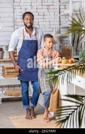 Black father and his son kid cooking fresh vegetables on kitchen at home. African dad and boy child preparing together. Stock Photo