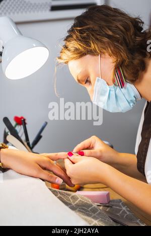 Manicurist with safety measures and face mask working on client's nails at manicure and pedicure salon after reopening of the pandemic. Stock Photo