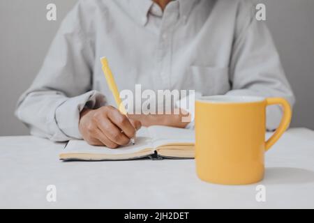 Close-up of a hand writing a note in a notebook  and a cup Stock Photo