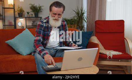 Happy smiling senior grandfather man accounting payments and calculating monthly expenses bank bills, analysing paperwork at home room couch. Elderly freelancer working online remote job on laptop Stock Photo