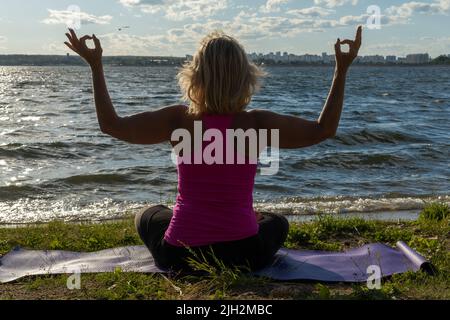 An elderly woman sitting in the lotus position and raising her hands up, on a mat in the park and looking at the lake, a view from the back. Meditatio Stock Photo