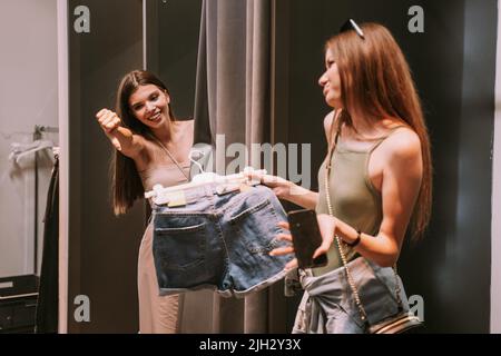 Two pretty best friends standing in the changeing room. One of the showing the shorts the other one disliking them Stock Photo