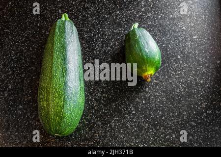 Green healthy and sick zucchini on a black table surface. Vegetable disease late blight, underdevelopment of fruits. Stock Photo