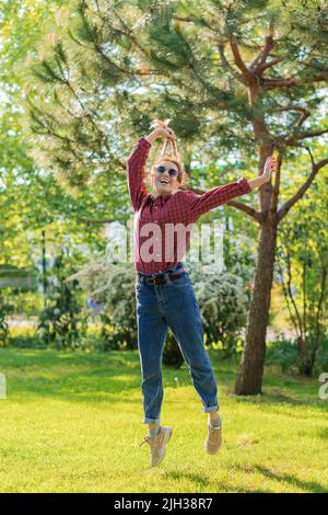 Funny woman lifts herself by her pigtails off the ground in green sunny park Stock Photo