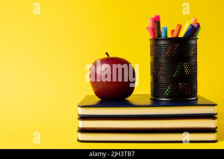 Composition of stack of books, felt tip pens in container and apple on yellow background Stock Photo
