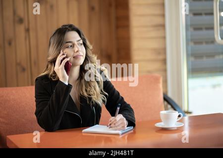 Young beautiful girl makes a phone call in a cafe, writes notes in a notebook. Student learning online. Freelancer working online. Stock Photo
