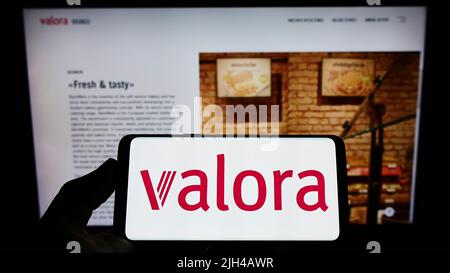 Person holding smartphone with logo of Swiss retail company Valora Holding AG on screen in front of website. Focus on phone display. Stock Photo