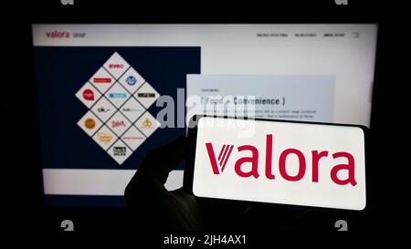 Person holding cellphone with logo of Swiss retail company Valora Holding AG on screen in front of business webpage. Focus on phone display. Stock Photo