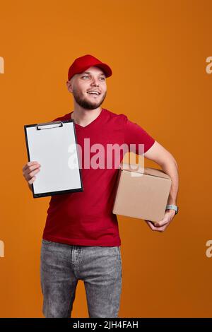 Delivery man in red cap, t-shirt holding clipboard and empty cardboard box isolated on orange background Stock Photo