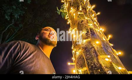 MIddle age bearded asian man standing near the led light bulb tree in the park. Low angle view. Stock Photo